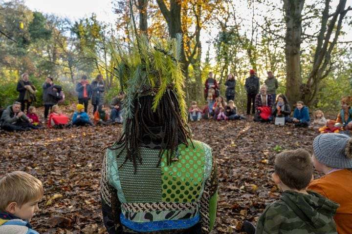 Children sat in a circle in the woods while performer is sat with back to the camera in a green head-dress.