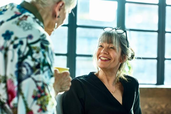 Woman in her 50s with glasses on head and grey hair, looking up with happy expression, listening to senior woman at work,
