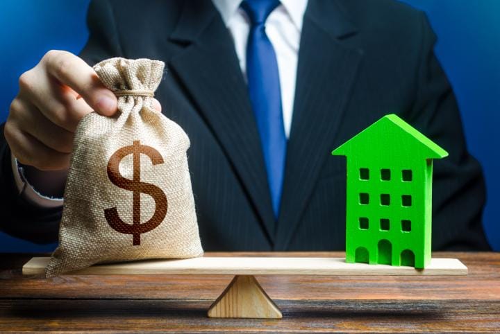 Businessman puts dollar money bag on scaled opposite a small painted wooden green house.