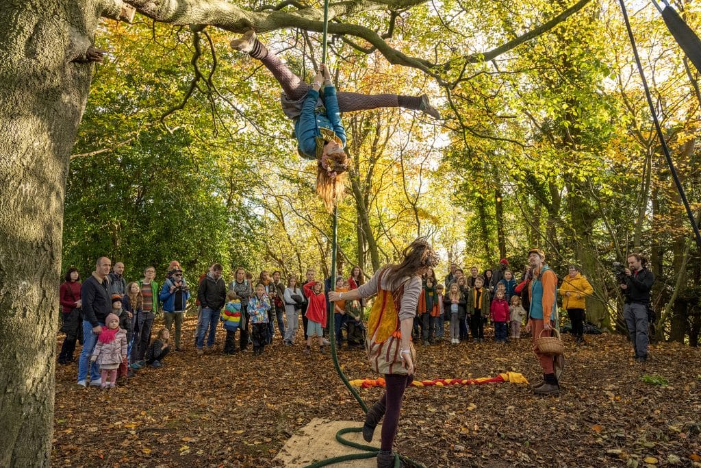 Performers hangs upside down on a from from a tree branch while school children parents watch.