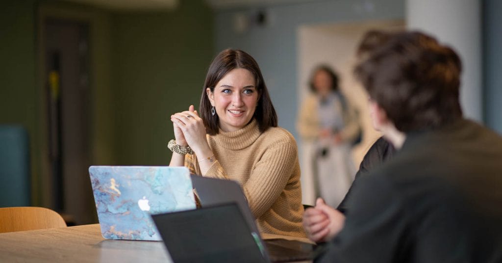 Photograph of three people consulting or discussing a topic while they use their laptops for reference. One other person can be seen entering the room in the background.