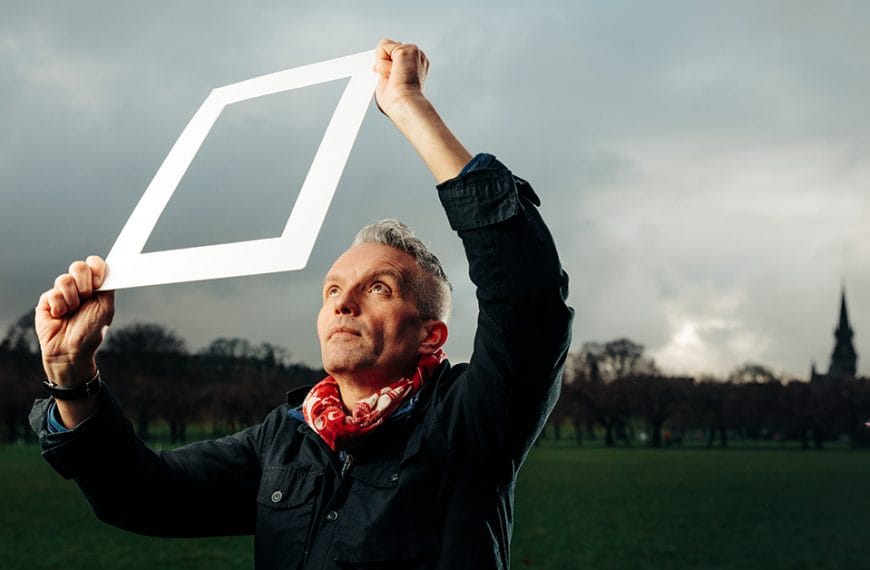 Professor Hayden Lorimer looking through a white cardboard frame at the sky with grey clouds and trees in the background.