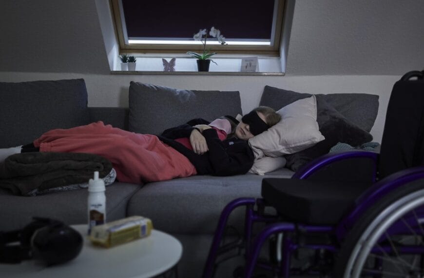Young girl with ME lies on sofa in a darkened room wearing an eye mask, with a wheelchair in the foreground