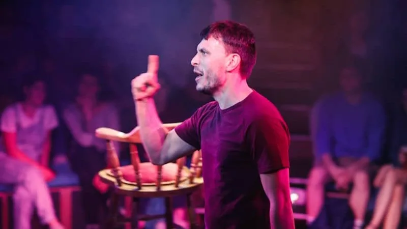 Actor Richard Gadd in a production image from his 2019 production Baby Reindeer, where he is pictured performing in an auditorium with a blurred audience in the background. 