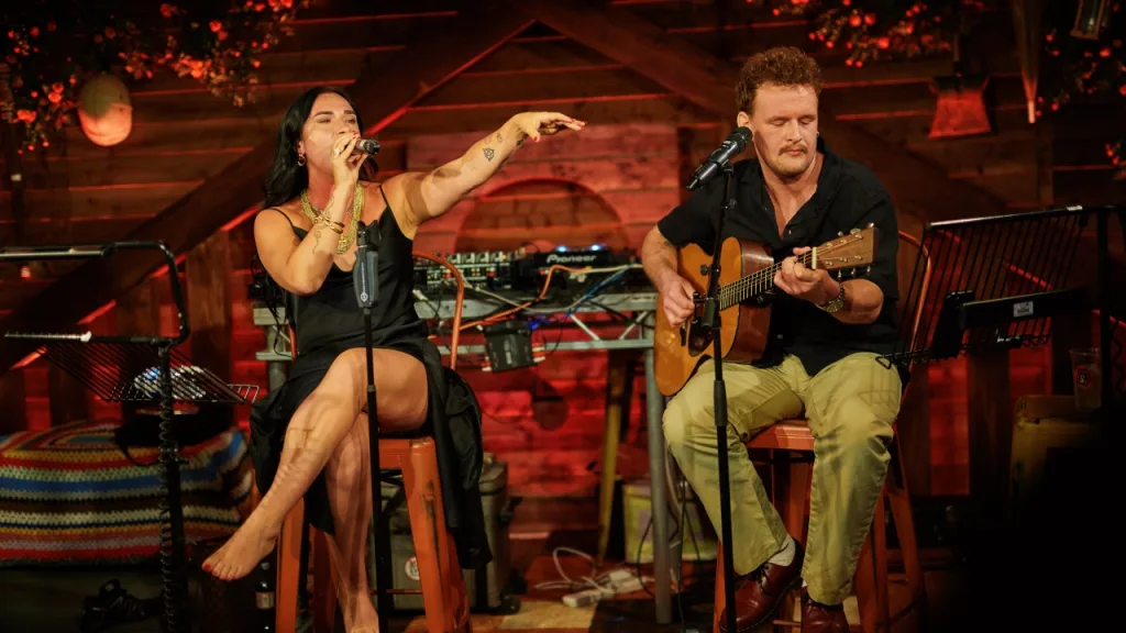 A musical duo perform on the Shedinburgh stage, with a man playing a guitar and a woman singing. 