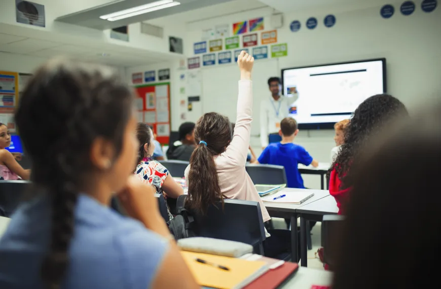 High school student raising hand facing a teacher in a classroom, asking a question during lesson.