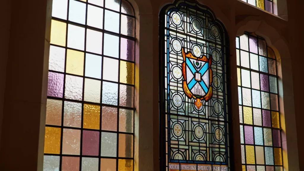 An interior view of light streaming through original stained-glass windows featuring the University crest, which have now been restored, helping to better insulate the building.