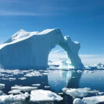 A large iceberg with pieces of ice floating in the ocean in the foreground and blue sky in the background