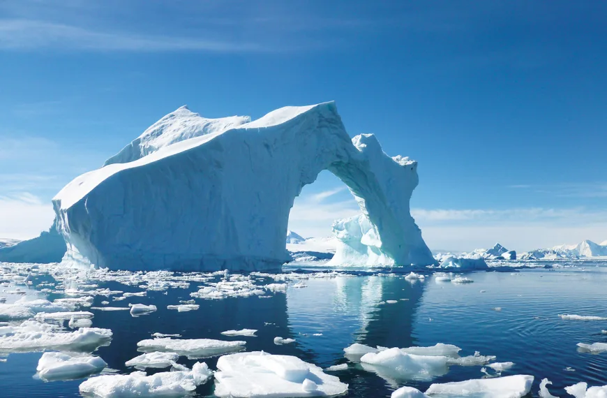 A large iceberg with pieces of ice floating in the ocean in the foreground and blue sky in the background