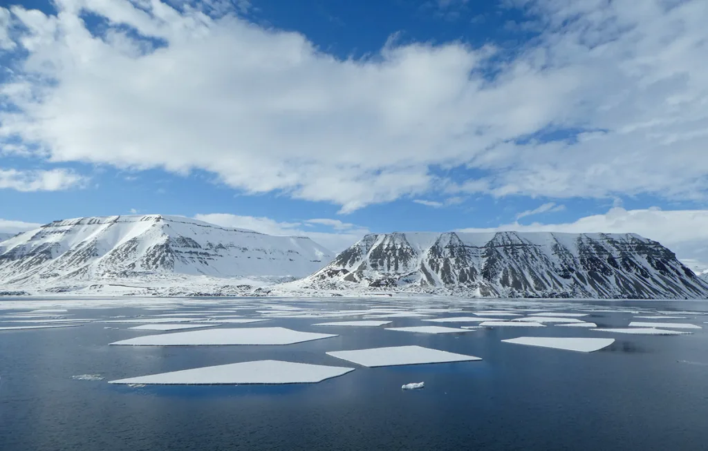 Ice floating on the ocean in the foreground with snowcapped mountains in the background with clouds across the blue sky