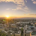 Panoramic image of Edinburgh, with the sun shining just above the urban skyline.