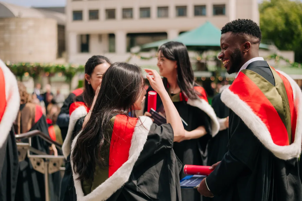Students wearing graduation robes with a red hood and white fur lining chat in Bristo Square at the University's Summer 2024 Graduations.