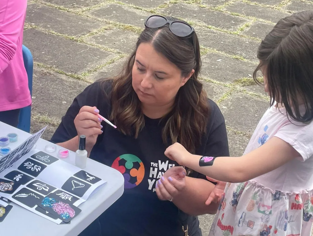 Sabrina helping a young person apply a transfer tattoo at a family fun day event at The Why Project