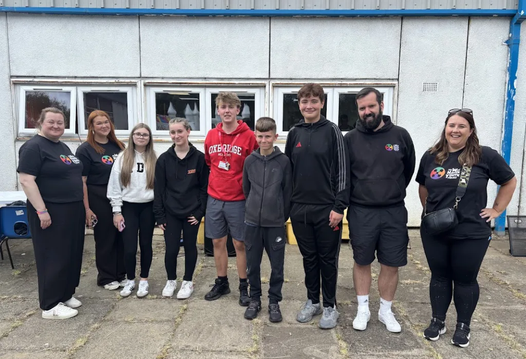 Sabrina (end right) and cofounder Kevin (next to Sabrina) stand in a line outside with a building in the background facing the camera with other helpers and young people involved in their youth work project