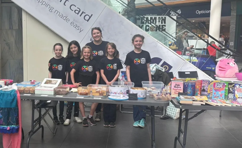 Sabrina with young people behind tables laid out with cakes, books and toys at a spring fayre to raise funds for their youth work.