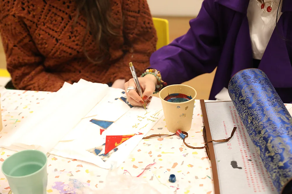 A close up of a table during one of artist Jiaya Chen's workshops showing her hand holding a pen  writing on paper with a pot of ink and a scroll of script and a blue patterned roll of paper in the foreground.