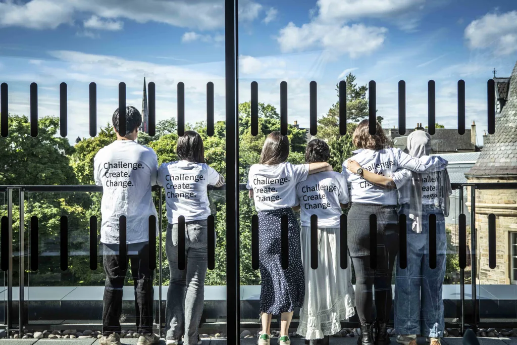 This photograph shows a group of six students staring out across the rooftop terrace of Edinburgh Futures Institute. They are relaxing and enjoying the view of blue sky, trees and rooftops of Edinburgh. Each of them wears a T shirt with the words 'Challenge. Create. Change.' on the back of the shirt. These are typical words to describe the work that happens in the Institute on a typical day for our students and staff.