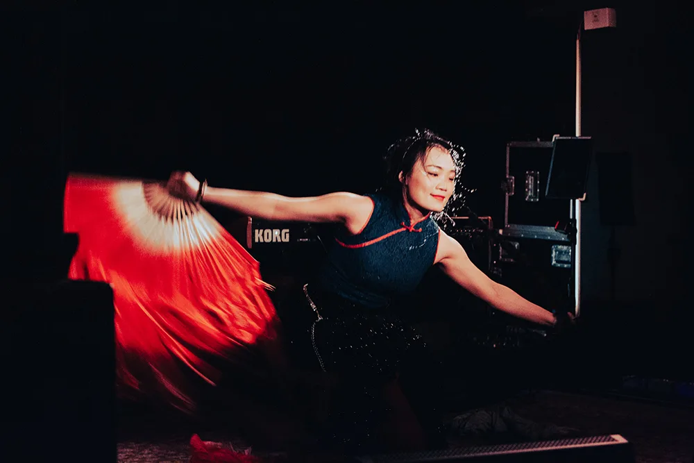 Performance artist Jiaya Chen on stage moving with her arms outstretched holding a large red fan.