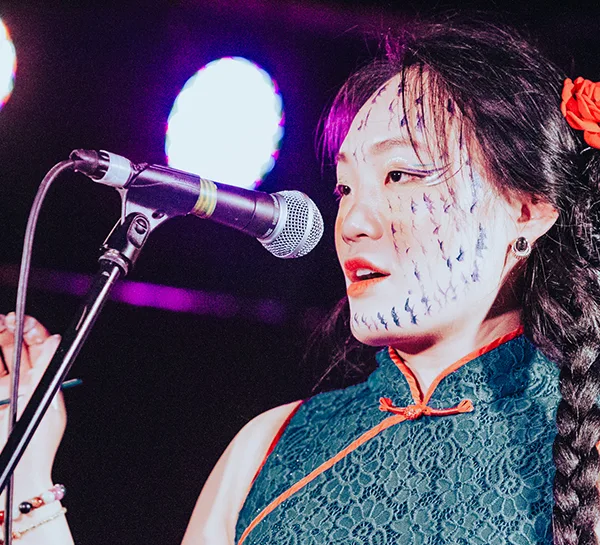 Performance artist Jiaya Chen on stage speaking into a microphone and holding a calligraphy paint brush