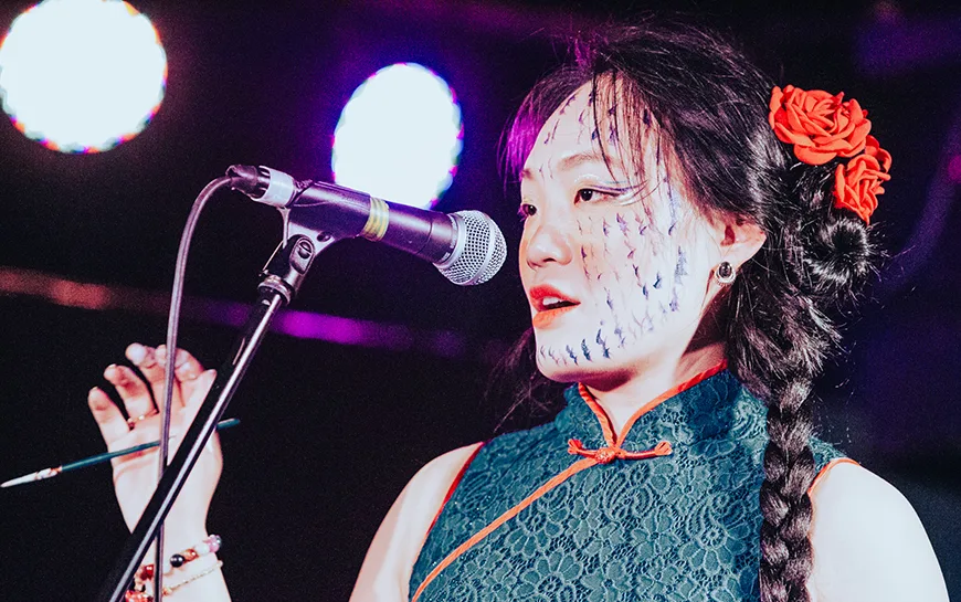 Performance artist Jiaya Chen on stage speaking into a microphone and holding a calligraphy paint brush