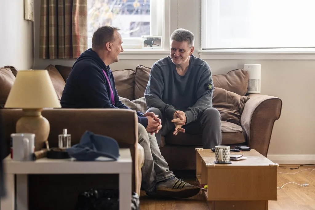 Two people supported by Simon Community Scotland sit on a sofa chatting.