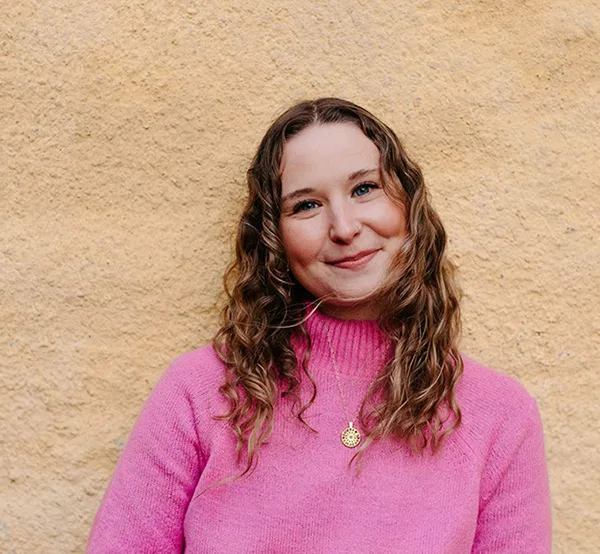 Emma Ahlert facing the camera smiling wearing a pink jumper with pale wall behind