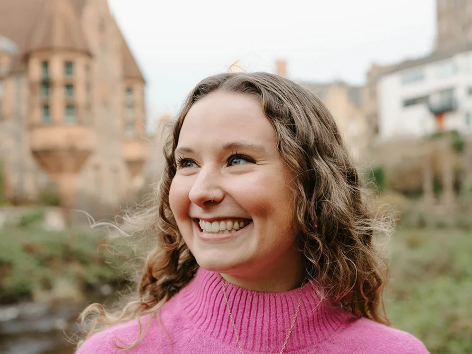 Emma Ahlert looking off camera to the left smiling wearing a pink jumper. Buildings and greenery are soft focus in the background.